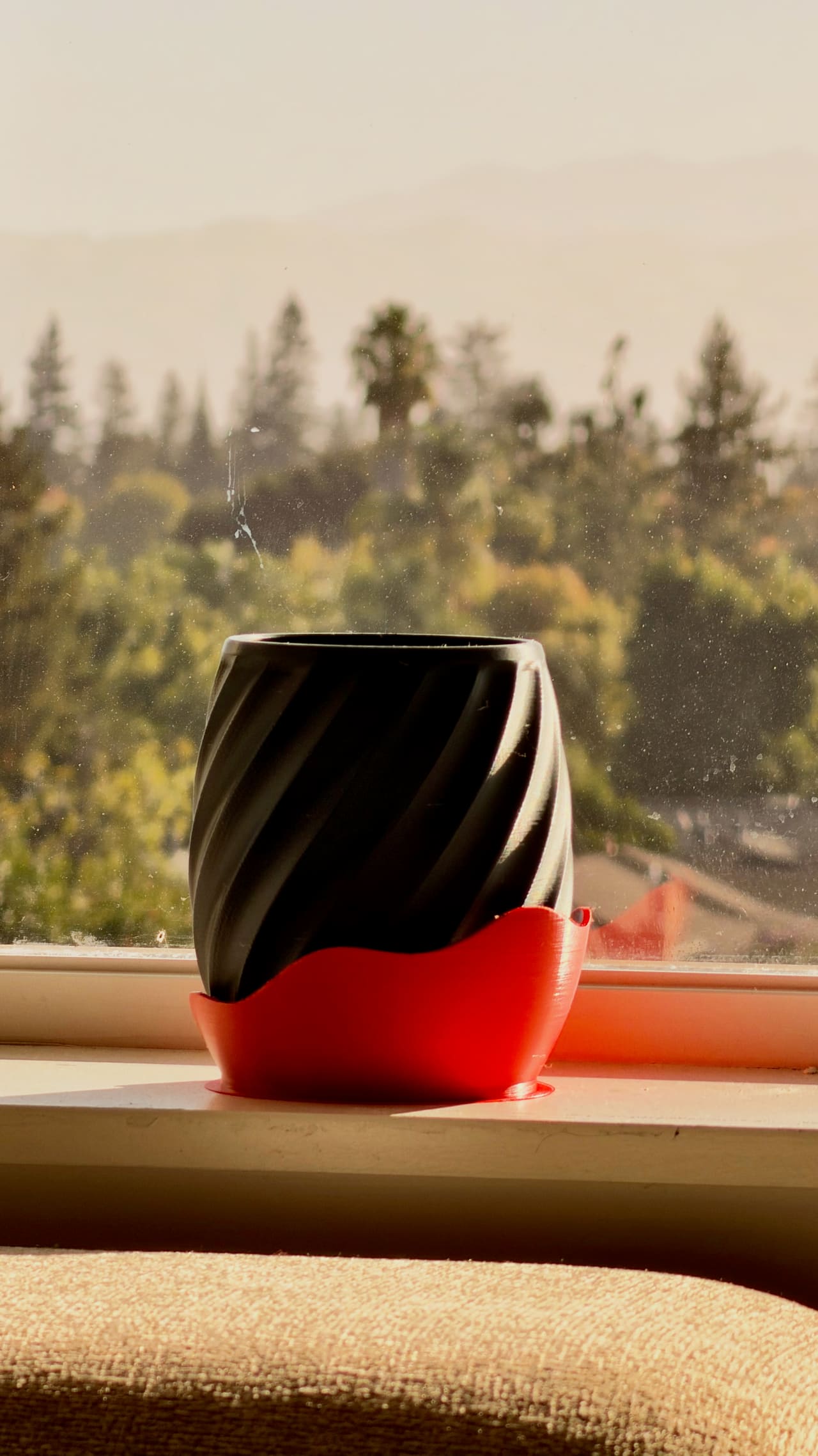 A black 3D-printed pot with a twisted surface, sitting in a red saucer with a wavy rim on a sunny windowsill